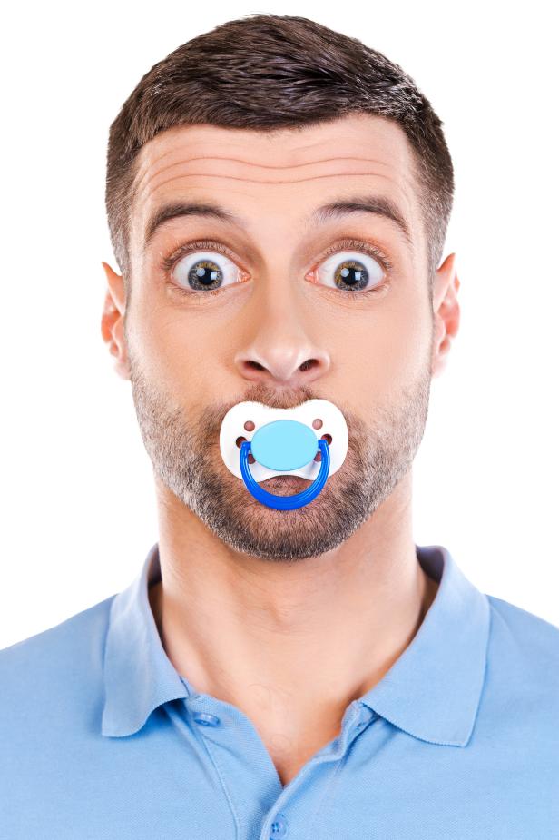 Funny young man with big eyes and pacifier in his mouth staring at camera while standing against white background