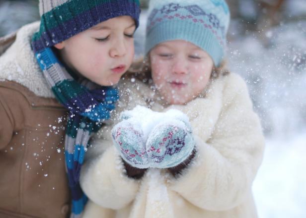 A young girl dressed in winter clothes is holding a handful of snow. The girl and her brother are blowing the snow out of her hands. The focus is on the hands and the snow.