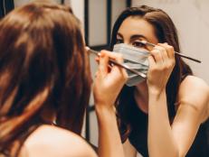 selective focus of young woman in medical mask and black dress applying eye shadow in bathroom