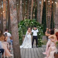 Happy exited newlyweds stands in the middle of wooden stage under the green floral arch. A lot of guests at the outdoor reception greet them and make pictures. Pine wood is on the background