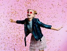 Female teenager wearing black leather jacket, pink tulle skirt and heart shaped sunglasses dancing among gold confetti. Studio shot on pink background.
