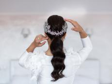 Fiancee with dark long hair turned her back to the camera. Hairstyle decorated with silver floral barrette looks amazing on the light background and pure morning light. Bride touches her head tenderly