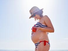 Pregnant woman putting on sunscreen on the beach with the sea in the background, she is wearing sunglasses and a hat
