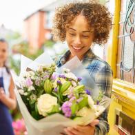 A young woman stands at the front door of her home and reads the card from a big bouquet of flowers . Behind her is the delivery girl who has just dropped them off. The shallow focus is on the bouquet blurring off to the people.