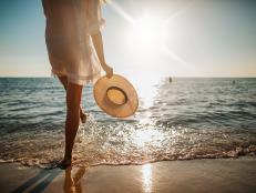 Close-up of young woman in white sun dress and with hat in hand walking alone on sandy beach at summer sunset, splashing water in sea shallow