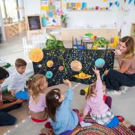 Multiracial group of small children with teacher sitting on floor and learning about space together at kindergarten.