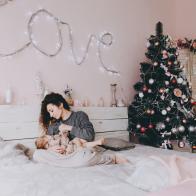 A young curly-haired mother in a gray sweater is breast-feeding her daughter on a trendy pink background in the Christmas and New Year's interior. Holiday. Family concept. (A young curly-haired mother in a gray sweater is breast-feeding her daughter o