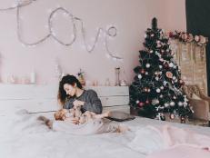 A young curly-haired mother in a gray sweater is breast-feeding her daughter on a trendy pink background in the Christmas and New Year's interior. Holiday. Family concept. (A young curly-haired mother in a gray sweater is breast-feeding her daughter o