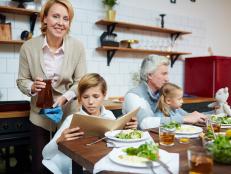 Cute boy reading book while sitting by served table with his sister and grandparents near by