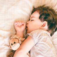 Boy sleeping on bed holding a soft toy by his side, against a soft cream background.