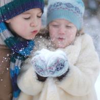 A young girl dressed in winter clothes is holding a handful of snow. The girl and her brother are blowing the snow out of her hands. The focus is on the hands and the snow.