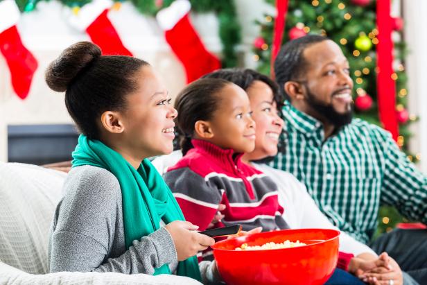 Happy African American couple and their teenage daughter and elementary age son watch a Christmas movie together during the Christmas season. The girl is holding a bowl of popcorn and the remote control. They are all smiling while watching the movie. A Christmas tree, stockings and a fireplace are in the background.