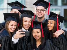 Multiracial group of six friends taking a selfie at graduation, in their black caps and gowns.
