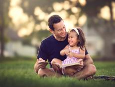 A smiling middle aged dad reads a silly story to his laughing 2 year old daughter at the park in front of his home.