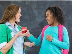 Adorable African American schoolgirl gives female teacher a apple on the first day of school. They are standing in front of a chalkboard.