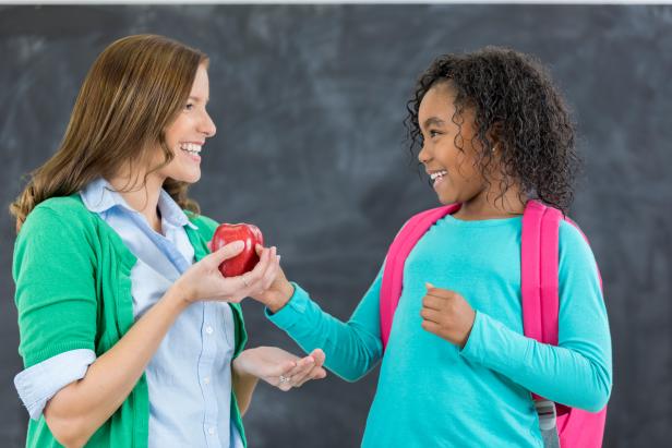 Adorable African American schoolgirl gives female teacher a apple on the first day of school. They are standing in front of a chalkboard.