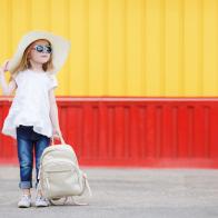 Beautiful little girl 6-8 years old,in a large straw hat from the sun,dressed in a short white dress without sleeves and blue jeans,with long blonde wavy hair and wears dark sunglasses in blue frame,posing standing near the school with a white backpack