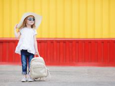 Beautiful little girl 6-8 years old,in a large straw hat from the sun,dressed in a short white dress without sleeves and blue jeans,with long blonde wavy hair and wears dark sunglasses in blue frame,posing standing near the school with a white backpack