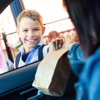 Young boy takes lunch from mother in carpool line. His mom hands him his brown paper bag lunch as he smiles at her. She is dropping him off at his elementary school.