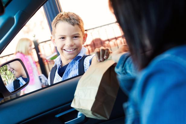 Young boy takes lunch from mother in carpool line. His mom hands him his brown paper bag lunch as he smiles at her. She is dropping him off at his elementary school.