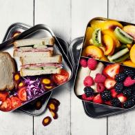 Two lunch boxes with sandwiches, veggies and fruits on white background.