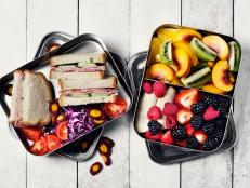Two lunch boxes with sandwiches, veggies and fruits on white background.