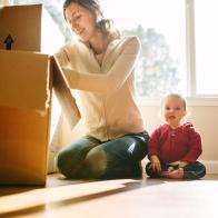 A mother unpacks cardboard moving boxes with her baby daughter sitting next to her with a big smile on her face.  Bright sun light shines in through the large bay windows in the living room.  Horizontal image.