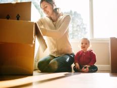 A mother unpacks cardboard moving boxes with her baby daughter sitting next to her with a big smile on her face.  Bright sun light shines in through the large bay windows in the living room.  Horizontal image.
