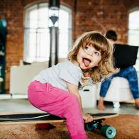 A toddler playing on a skateboard indoors while her mother works on a laptop.