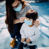 Young mother putting on surgical mask for little daughter in the playground to prevent the spread of cold and flu and viruses