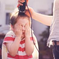 A young boy having his hair cut by his mother using hair clippers in their back garden.