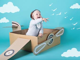 A baby boy is sitting inside a cardboard box made into an aeroplane. He is wearing a flight helmet and pointing at some birds and clouds.