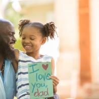 Happy Father's Day.  Little girl gives homemade card to her dad in front yard of family home.  African descent family.