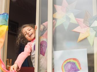 Preschool age little girl at her home window is waving her rainbow silk while quarantined. On the window you can see some rainbow artworks as a way to spread hope and cheers in these dark days.