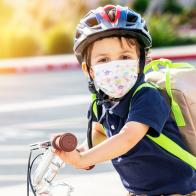 Caucasian Little boy riding a bicycle wearing a protective mask and a backpack looking at the camera