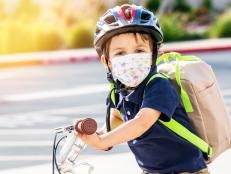 Caucasian Little boy riding a bicycle wearing a protective mask and a backpack looking at the camera
