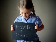 Four year old girl with plaits and blue check dress holds a handwritten chalkboard sign that says, "First Day of School".
