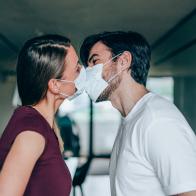 Beautiful happy young couple wearing protective face masks and kissing each other. Side view of young man and young woman in medical masks kissing while they are in home isolation during coronavirus/COVID-19 quarantine.