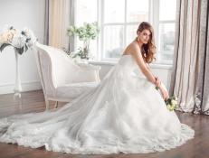Wedding. Bride in beautiful dress sitting on sofa indoors in white studio interior like at home. Trendy wedding style shot in full length. Young attractive caucasian brunette model like a bride against big window tender posing.