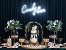 Big table covered with dark turquoise tablecloth is filled with dozens of desserts. Wedding cake muffins, mousses and macaroons are set on the stylish wooden plates. Dark curtain is on the background