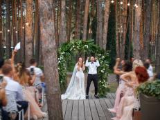 Happy exited newlyweds stands in the middle of wooden stage under the green floral arch. A lot of guests at the outdoor reception greet them and make pictures. Pine wood is on the background