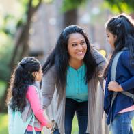 A mature Hispanic woman in her 40s standing with her two young daughters, 4 and 6 years old, holding their hands. The children are carrying backpacks on their way to school. The mother is bending down, looking at the older girl, smiling and offering words of encouragement.