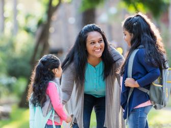 A mature Hispanic woman in her 40s standing with her two young daughters, 4 and 6 years old, holding their hands. The children are carrying backpacks on their way to school. The mother is bending down, looking at the older girl, smiling and offering words of encouragement.