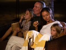 Family sitting on sofa enjoying movie night outdoor at home backyard together. Mother, father and children watching projector, TV, movies with popcorn in the evening