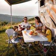 Happy family having fun while singing at picnic table by the camper trailer in nature. Man is playing a guitar.