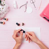 Female hands apply shellac during manicure procedure, flat lay. Woman painting her nails with gel nail polish near UV lamp, tools and bouquet, view from above.