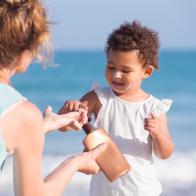2 years old little girl using sun protection cream for skin care. Grandmother showing little girl how to apply sunscreen. Grandmother and daughter enjoying summer holiday on sea beach together.