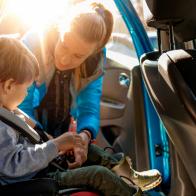 Young mother putting her little boy in the car seat, fastening seat belts.