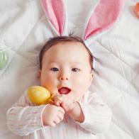 Cute adorable Asian baby wearing pink Easter bunny ears. Infant kid lying on bed with colored Easter eggs. Funny child celebrating traditional Christian holiday. Banner header for website.