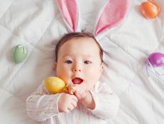 Cute adorable Asian baby wearing pink Easter bunny ears. Infant kid lying on bed with colored Easter eggs. Funny child celebrating traditional Christian holiday. Banner header for website.